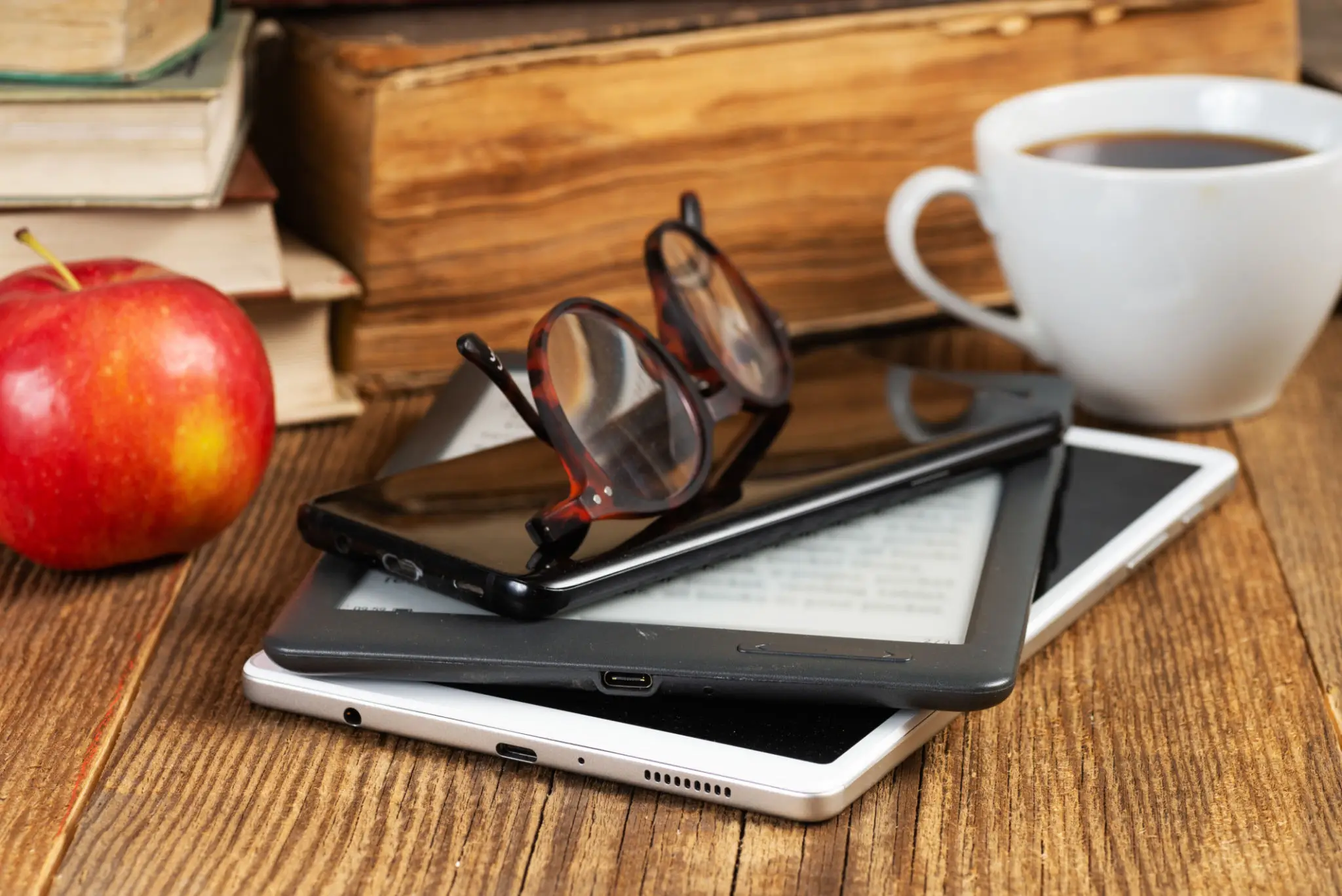 Stack of digital devices with glasses on top on a wooden table.